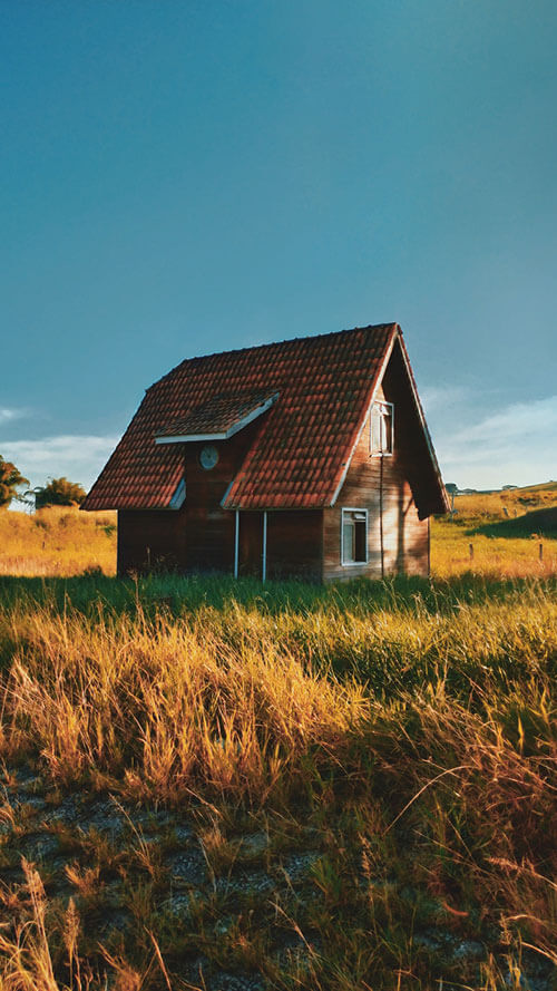 A small, two-story rustic wooden house with a red tiled roof standing in a golden grass field under a clear blue sky.